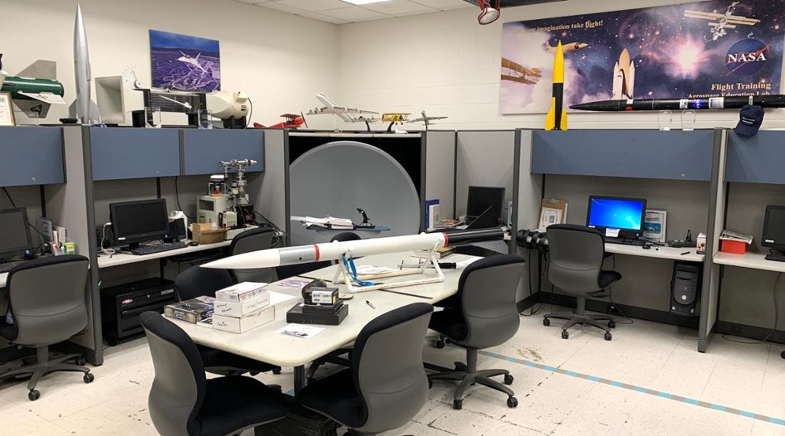 Lab with a table in the middle with a rocket on it. Desks and shelves filled with computers and other rockets and parts.