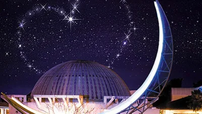 Night time view of imaginarium dome with a glowing moon sculpture in the foreground and heart shape twinkling in the stars